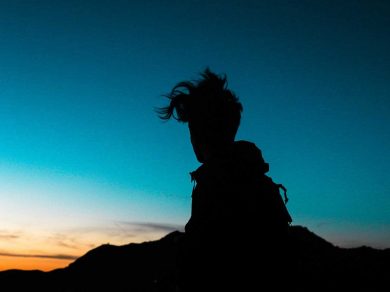 silhouette of young boy with wildly untidy hair sitting on rock with sunset behind